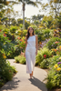 A woman wearing a white sleeveless top and matching white wide-leg pants walks along a garden path surrounded by lush tropical plants and colorful flowers.